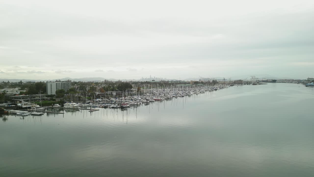 A cloudy aerial drone view of Mariner Square in Alameda, California, showcasing its bustling marina, docked boats, and scenic waterfront under soft golden sunlight.