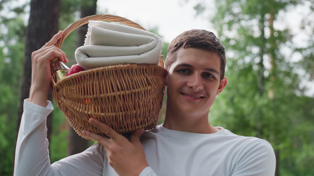 Portrét veselého mladého muže, který si s úsměvem zvedá košík na rameno a stojí u auta v zeleném lese, užívá si čerstvého vzduchu, slunečního světla a venkovní relaxace během letního dobrodružství.