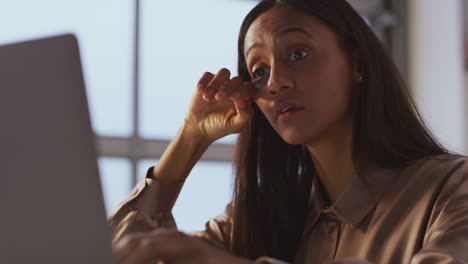 tired businesswoman working late using laptop at desk in office rubbing eyes and drinking coffee