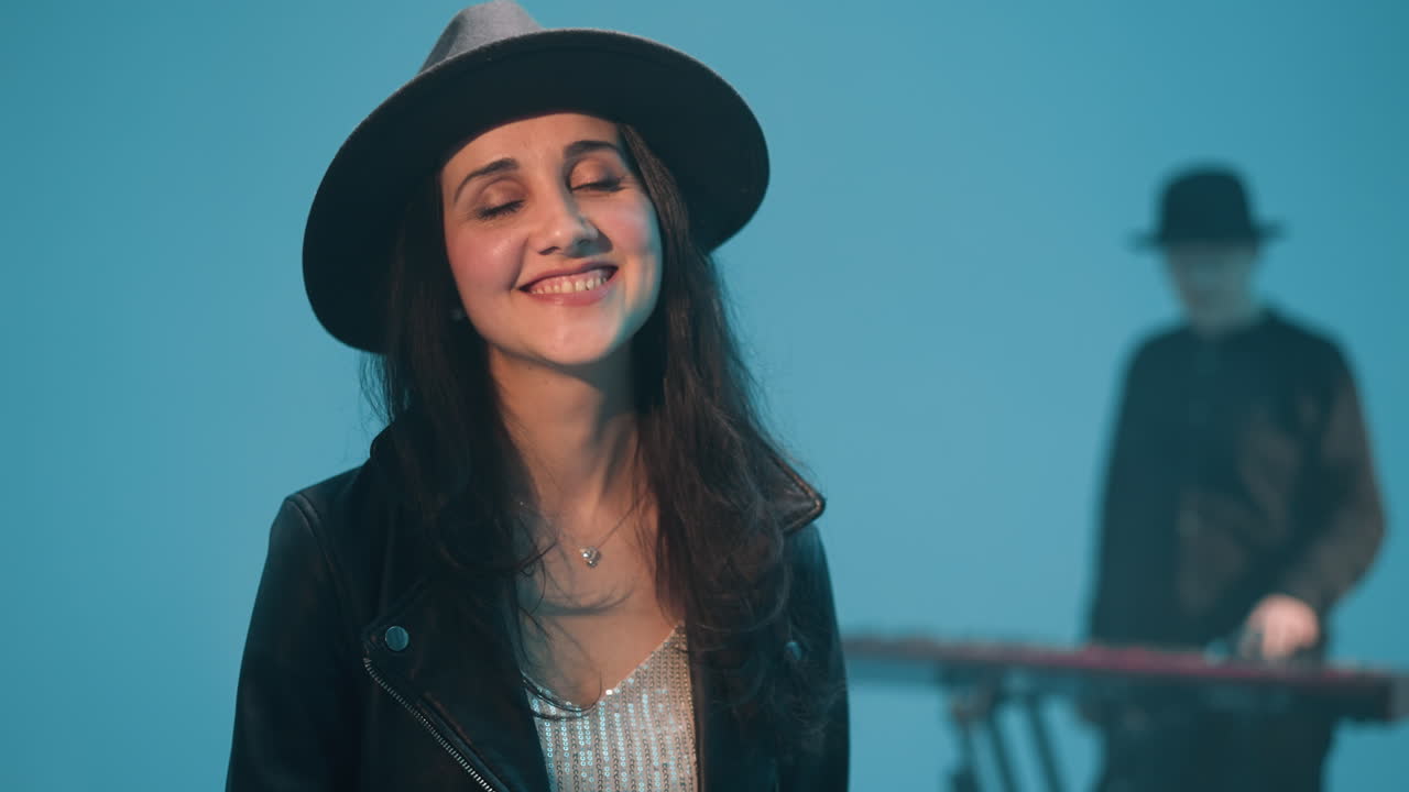 dama alegre con una chaqueta de cuero negra y sombrero sonriendo brillantemente mientras mira hacia arriba. en el fondo, un tecladista está borroso contra un telón de fondo azul, ambiente alegre de la escena