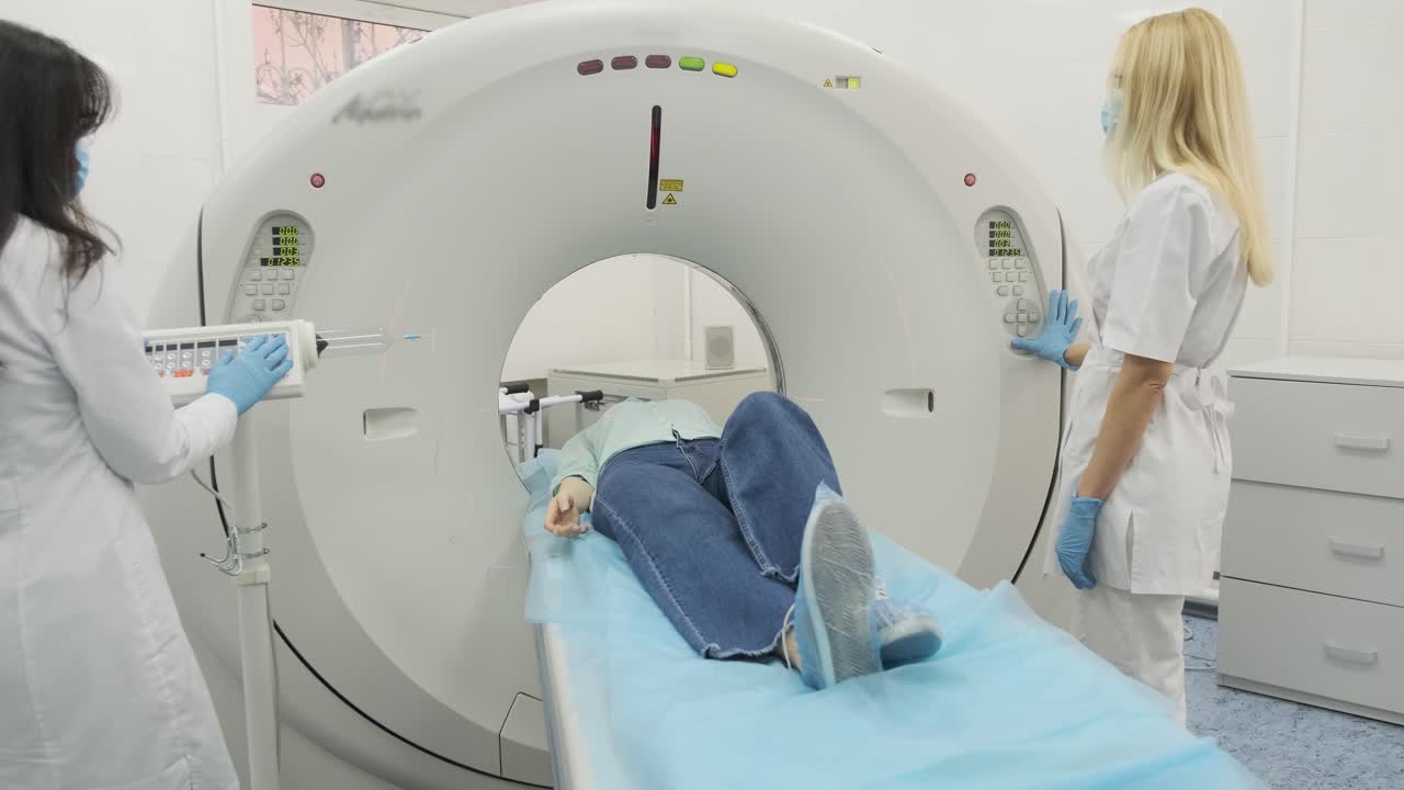 la paciente está siendo sometida a una tomografía computarizada o resonancia magnética bajo la supervisión de dos radiólogos calificados en una clínica médica moderna. la paciente está acostada en una mesa de tomografías computarizadas o mri, moviéndose fuera de la máquina