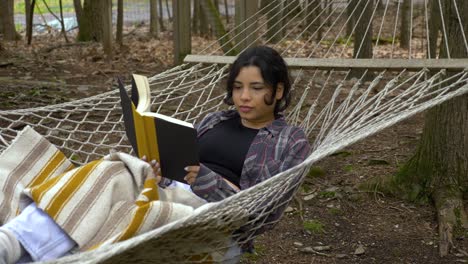 joven puertorriqueña cómoda leyendo un libro balanceándose en una hamaca en el bosque