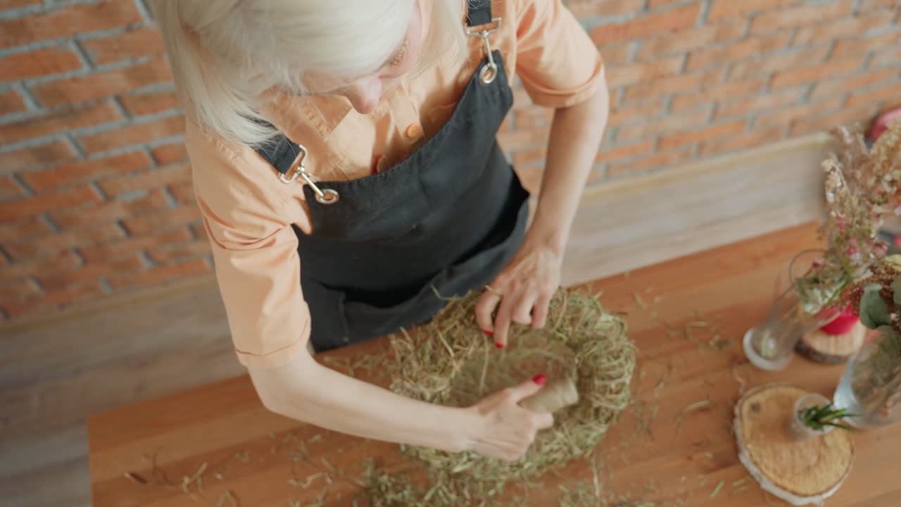 Woman florist in apron crafting straw wreath on wooden table, arranging dried grass with focused hands, creating handmade rustic decoration in cozy workspace emphasizing creative artisan process