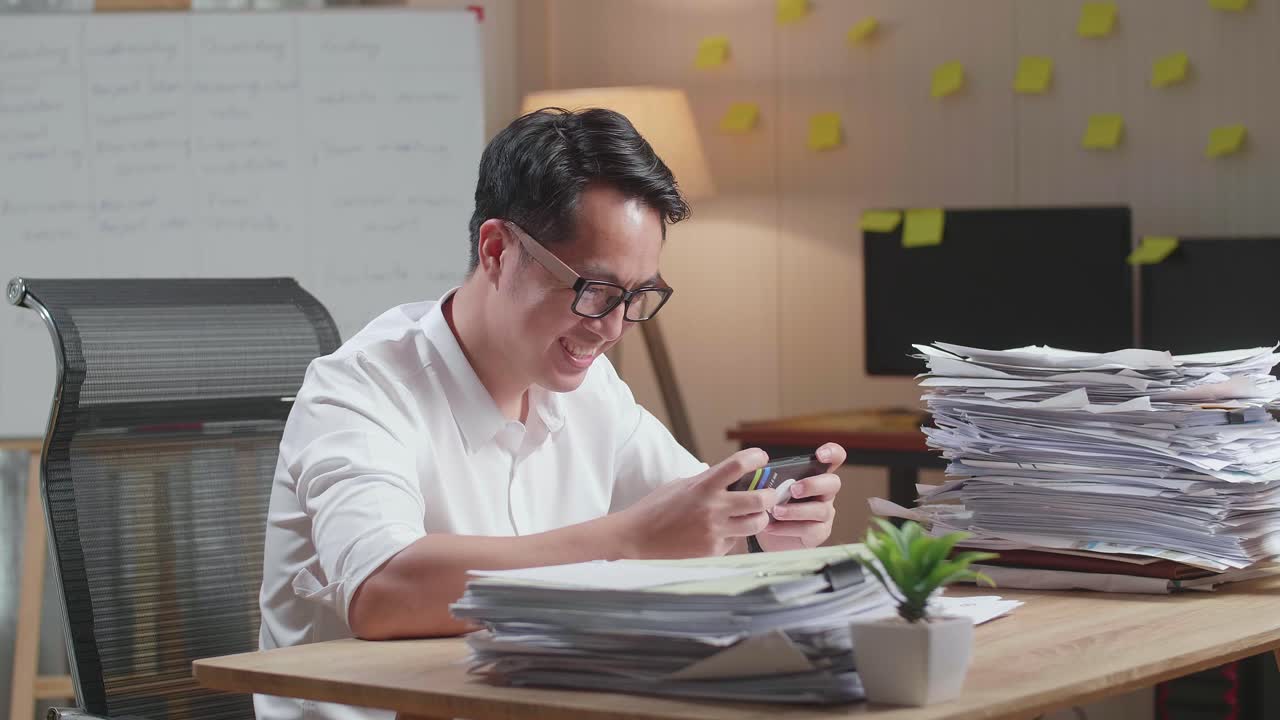 Asian Man Enjoys Playing Game On Smartphone After Working With Documents At The Office