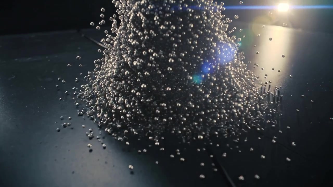 Cucitura che rilascia centinaia di sfere metalliche, formando un alto cono su un pannello di metallo scuro, luci che brillano