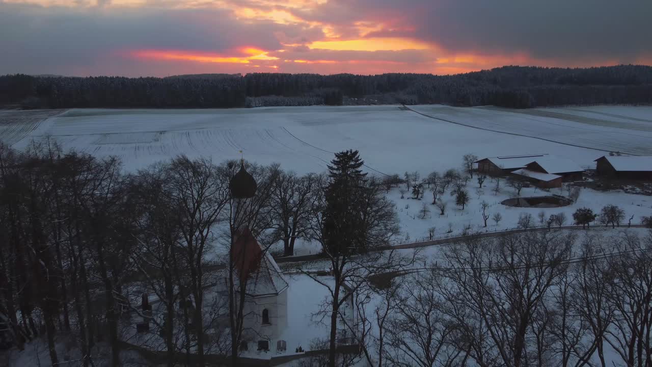 una antigua iglesia capilla con una cúpula redonda en una colina rodeada de árboles en invierno en baviera, alemania, con nieve en los campos vista desde arriba, imágenes aéreas de drones mientras se pone el sol con nubes rojas y cielo