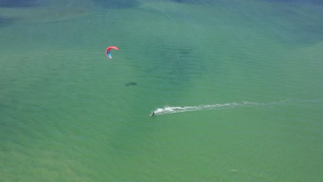 Cometa---Windsurfista-Se-Eleva-A-Través-De-Aguas-Turquesas,-Azules-Y-Verdes-Del-Océano-Frente-A-La-Costa-De-Fuzeta