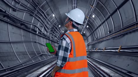 back view of asian male engineer with safety helmet working on a green screen smartphone and looking around while standing in underground subway tunnel