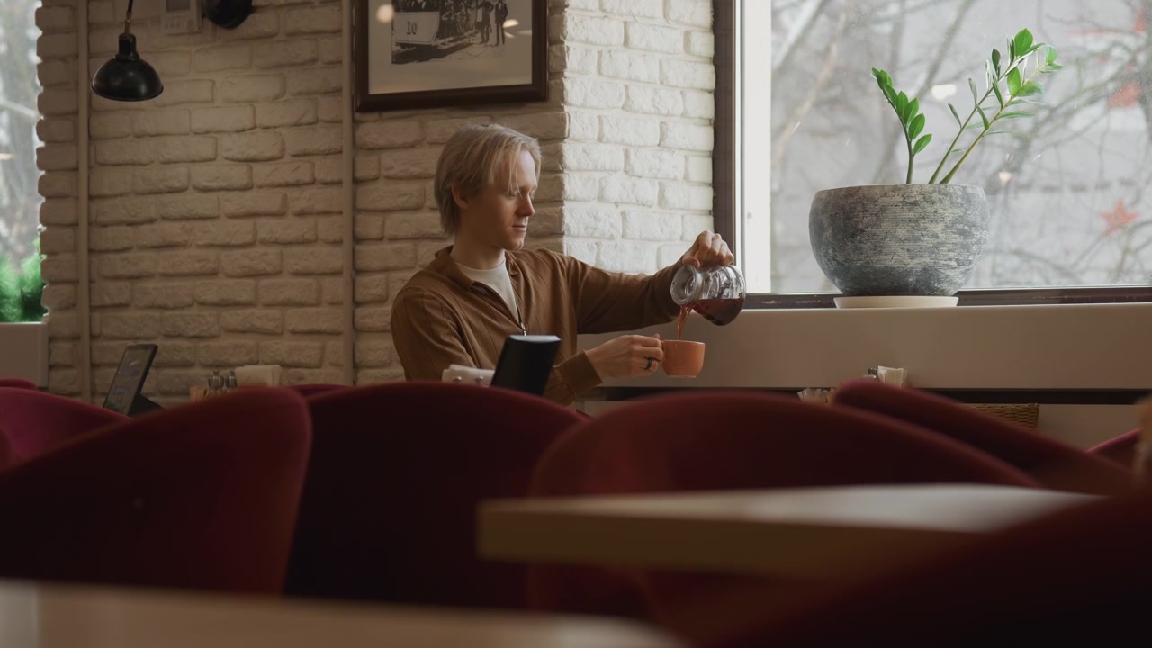 Close up of man sitting in empty cafe holding and pouring coffee into cup while looking out window. setting features white brick walls red chairs natural light from large windows framed