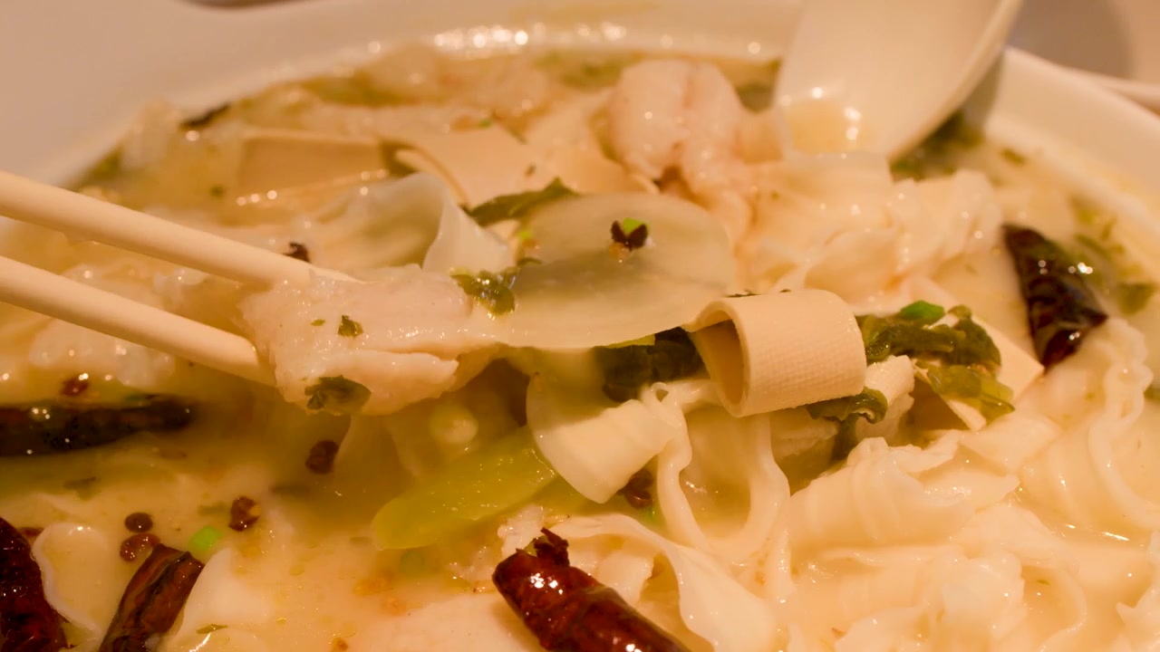 Macro di bacchette che prendono pesce da una zuppa di noodle piccante in una ciotola di ceramica.
