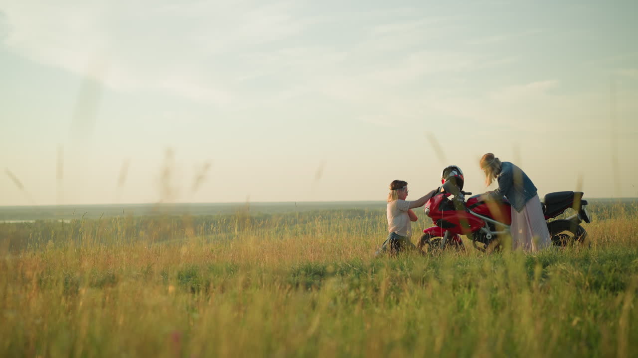두 여동생은 은 하늘 아래 잔디로 인 야외에서 빨간 모터사이클을 청소하는 것을 볼 수 있습니다. 하나는 크를 는 데 집중하고 다른 하나는 자전거의 다른 부분을 청소하기 위해 드려 있습니다.