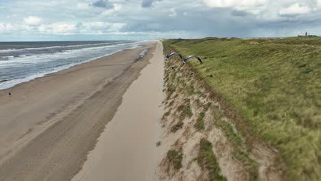 Cinematic-Aerial-Drone-Follow-Shot-of-Two-People-Paragliding-a-long-a-Beach-on-a-Moody-Cloudy-Sunny-Day