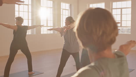 yoga class of healthy mature women practicing warrior pose enjoying morning physical fitness exercise workout in studio at sunrise