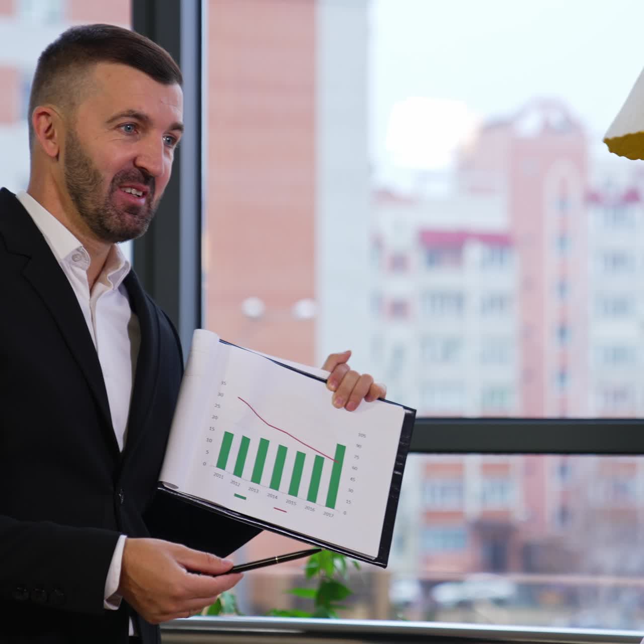Male mid-aged boss stands in front of his team and shows a document. Businessman explaining chart to his office staff at meeting