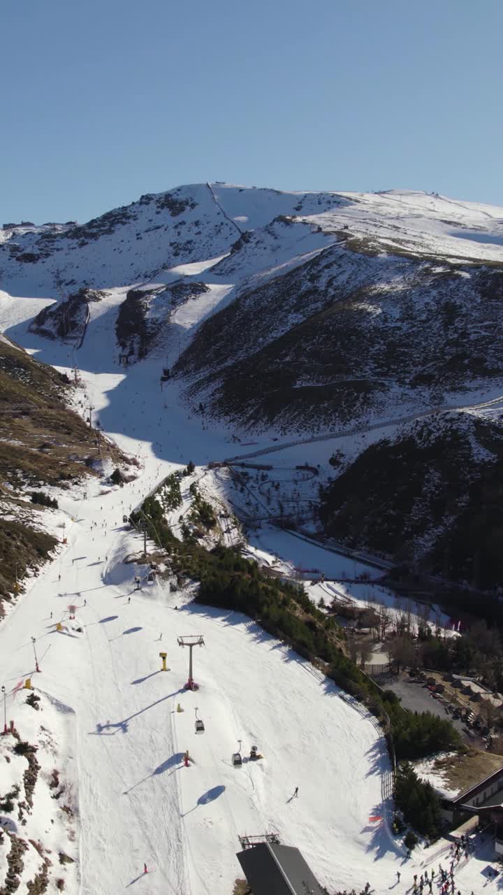 En drönare flyger över en bergsstad i Sierra Nevada, mot snöklädda toppar och skidspår i Andalusien. Vertikal flygbild.