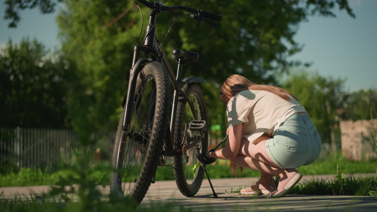 여성 자전거 타는 여성이 자전거 에 앉아서 은 날에 초록색 노즐로 타이어에 공기를 프합니다. 자전거는 초점에 서 있으며, 울타리, 나무 및 그림자 배경은 부드럽게 흐릿합니다.