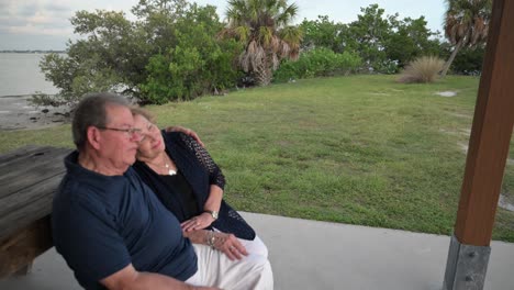 senior-citizen-married-couple-sit-on-a-park-bench-near-the-ocean-talking-and-enjoying-life