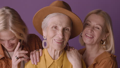 senior woman in hat and mustard shirt poses with two blonde mature women on purple background smiling at camera