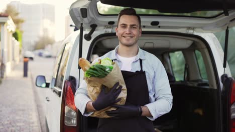 Glücklicher-Lieferarbeiter,-Der-Eine-Papiertüte-Mit-Lebensmitteln-In-Der-Hand-Hält,-In-Die-Kamera-Schaut-Und-Lächelt.-Nahaufnahme-Eines-Porträts-Eines-Positiven-Jungen-Mannes