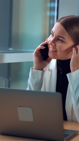 business lady having phone conversation sitting with laptop in front of her. woman talks and smiles looking at window. vertical video