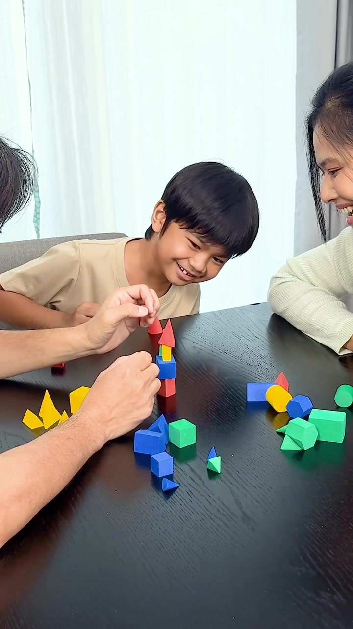 Un bambino sorridente e suo padre impilano blocchi geometrici colorati su un tavolo scuro all'interno.