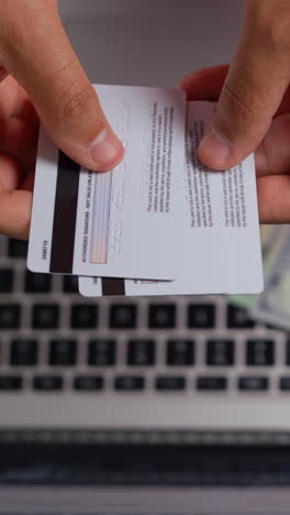 vertical video close-up of hands holding two credit cards over a laptop keyboard. soft indoor lighting highlights card details, perfect for mobile and social media use