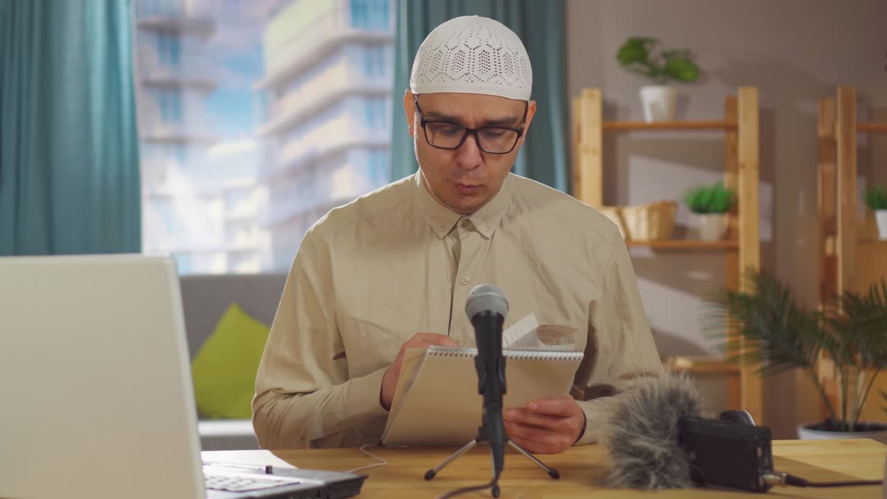 Portrait muslim man in a skullcap and wearing glasses records a podcast while sitting in front of a microphone in living room