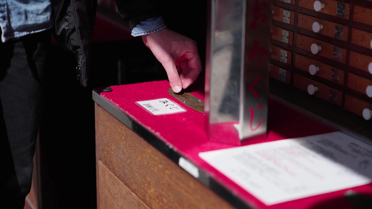Hand shaking cylindrical omikuji container on red velvet counter beside numbered wooden drawers shrine Tokyo Japan