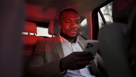 A-confident-man-with-Black-skin-in-a-brown-suit-with-a-short-haircut-and-beard-rides-in-the-interior-of-a-modern-car-and-works-on-a-white-smartphone