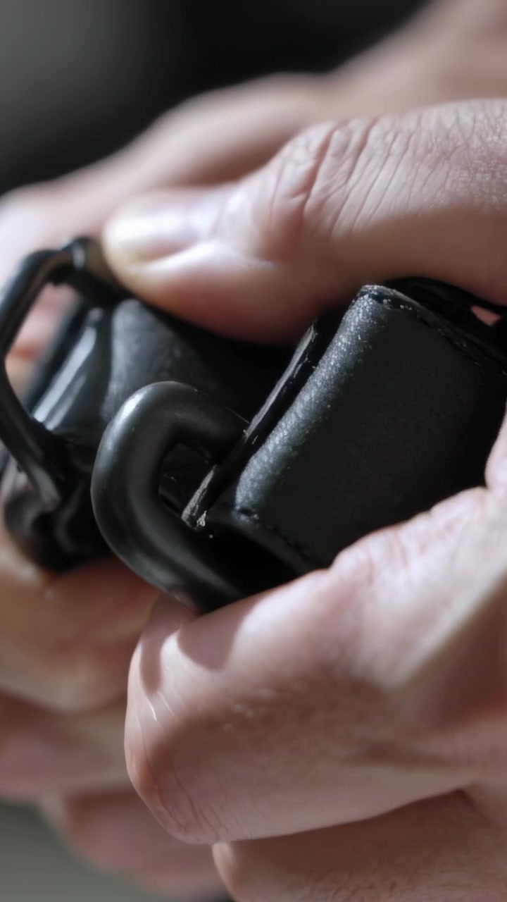Video vertical: Manos de adulto presionando el pulgar, ajustando una correa negra y una hebilla de metal en una habitación para un ajuste perfecto.