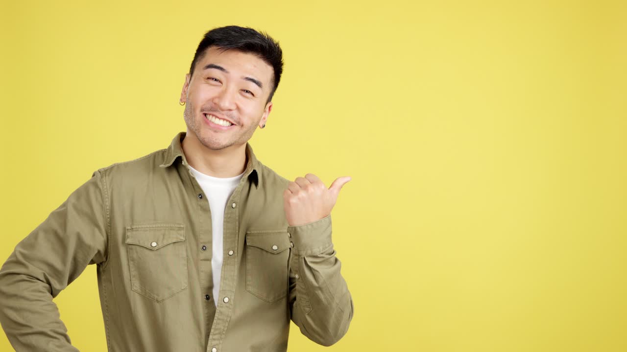 Smiling Asian Man Pointing with Thumb on Yellow Background
