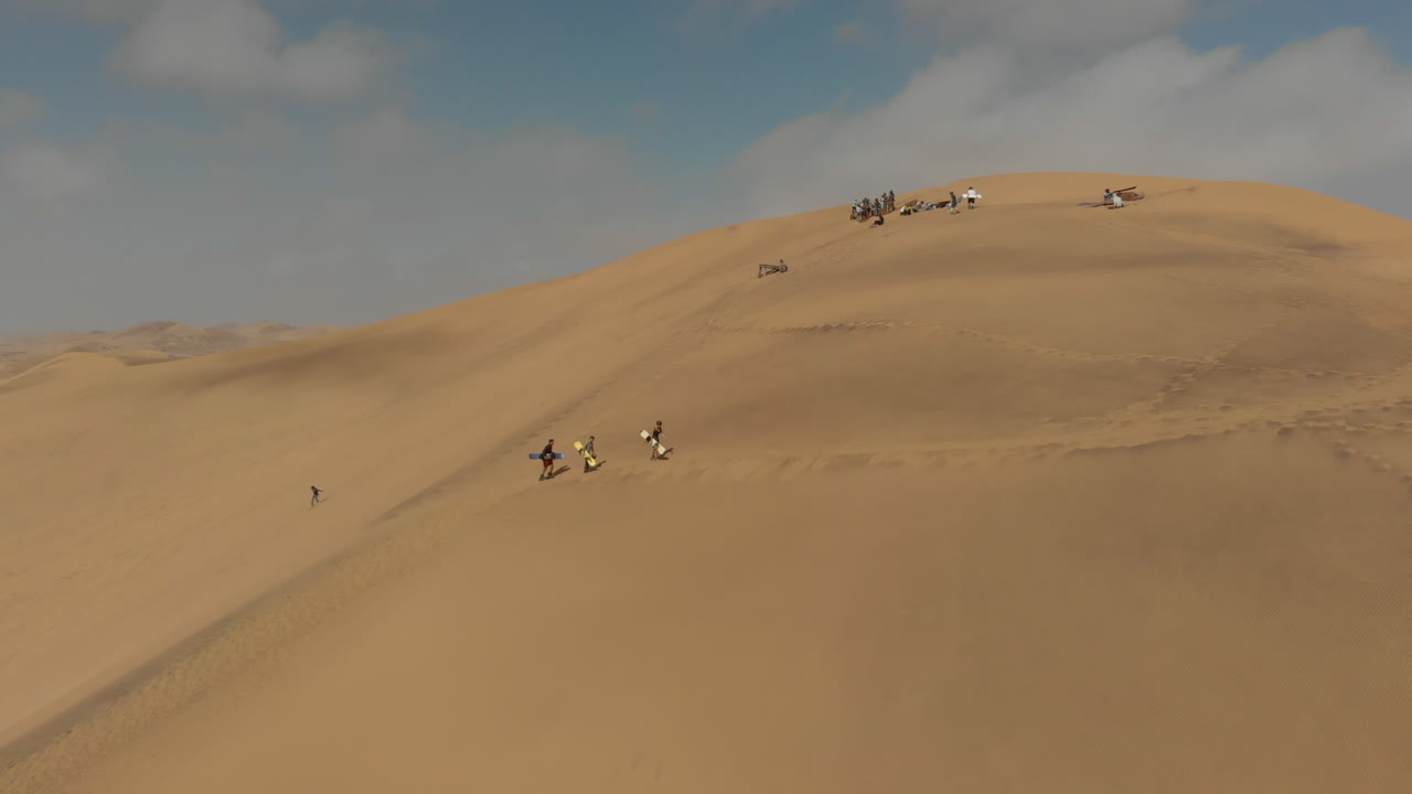 toma aérea de personas caminando por una duna de arena con tablas de arena