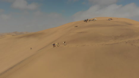 Toma-Aérea-De-Personas-Caminando-Por-Una-Duna-De-Arena-Con-Tablas-De-Arena