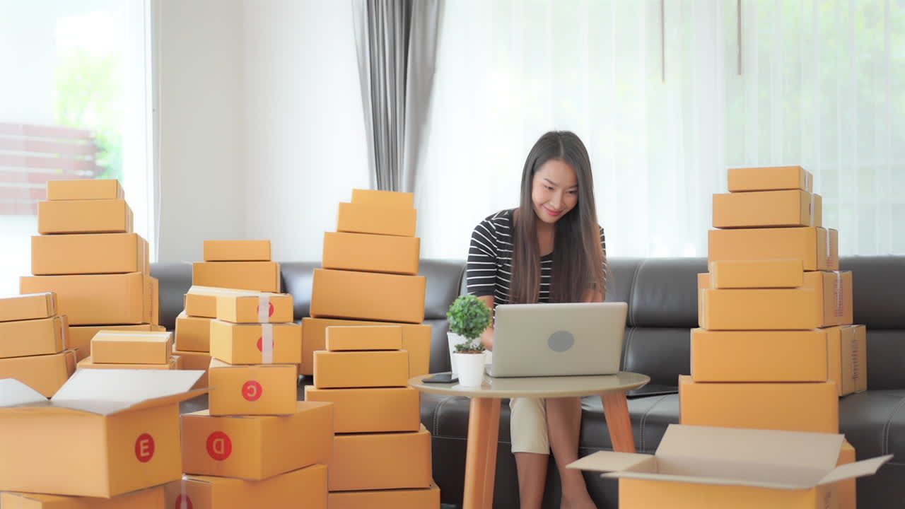 Working from home, a young woman surrounded by shipping boxes is buying inputting data into a laptop