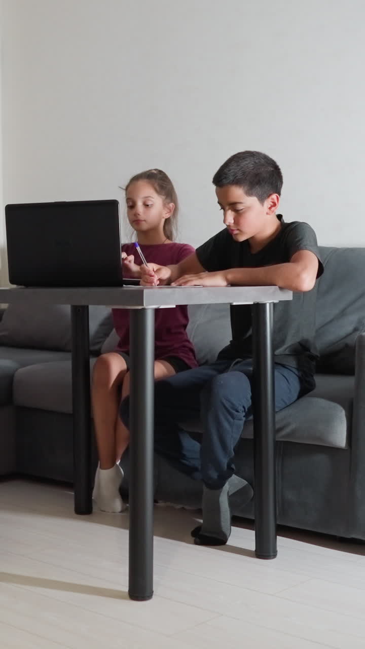 Hermanos haciendo los deberes juntos en el salón; el niño se concentra en escribir, mientras la niña usa el portátil; trabajando juntos en un momento de estudio tranquilo.