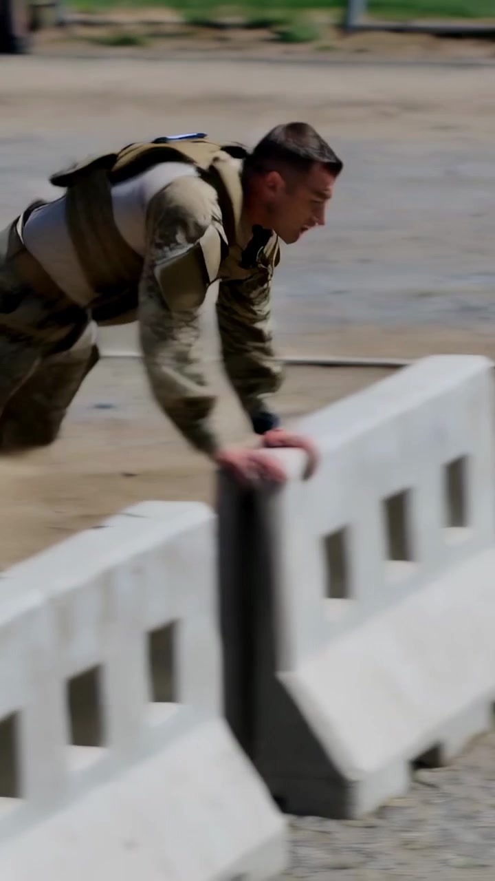 Vertical video: Running man entering left in camo wearing vest vaulting barriers at training yard
