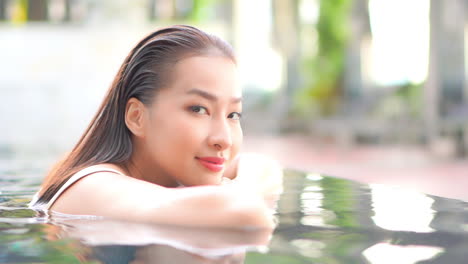 dreaming asian woman leaning head on the edge on infinity swimming pool and looking at camera smiling at sunset in thailand resort, face closeup slowmotion