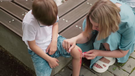 a mother gently squats and cleans her son's injured leg with a tissue while another person is partially visible in the background