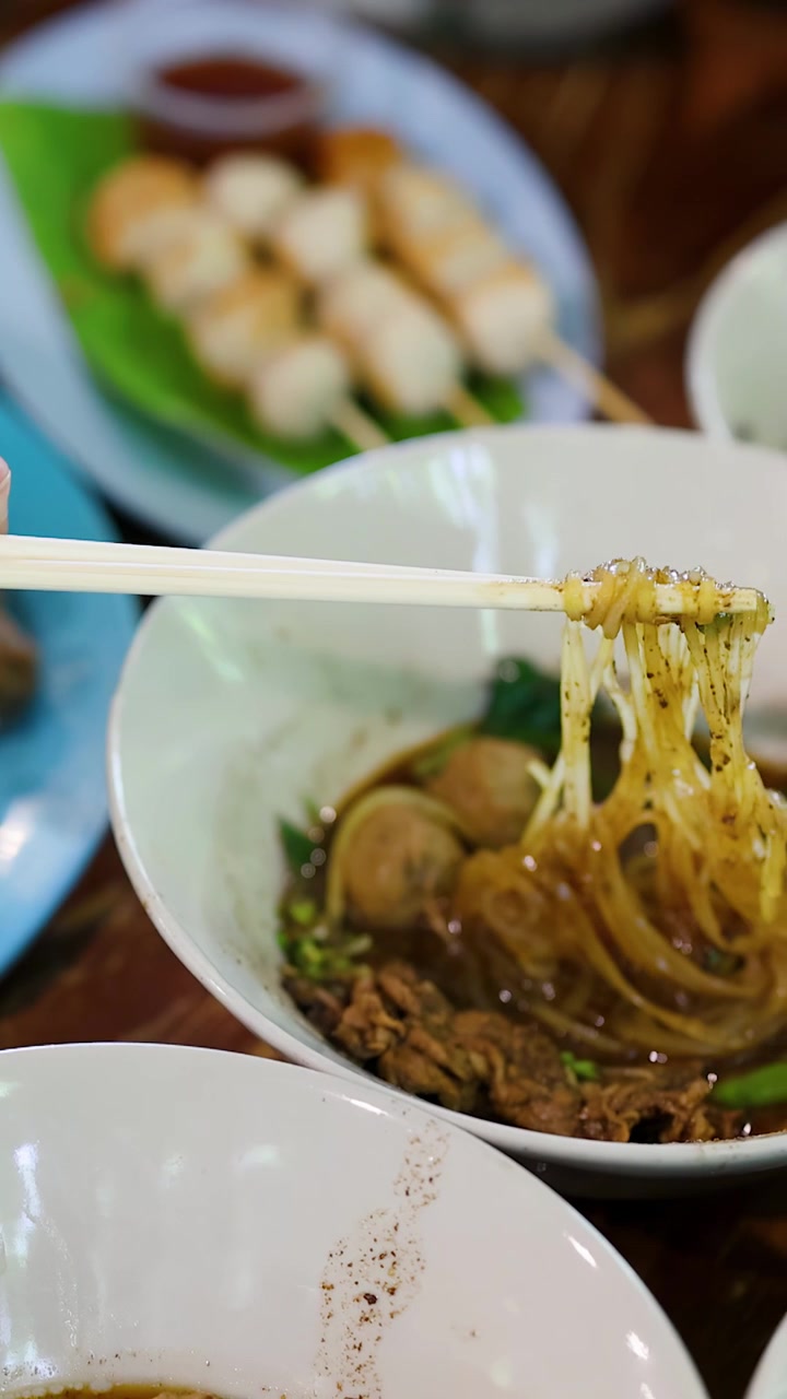 Close-up tangan menggunakan sumpit untuk mengangkat mi dari semangkuk sup bakso.