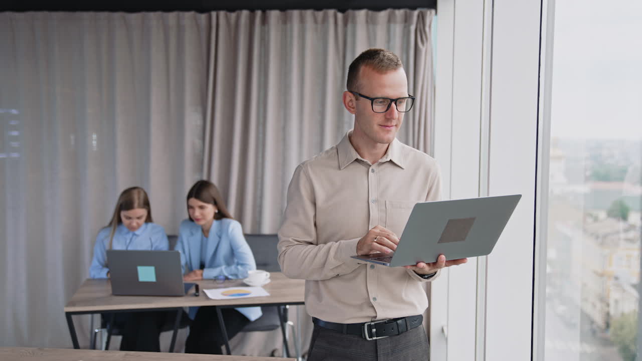 Mid-aged man in glasses working on laptop standing near the window. Female colleagues sitting and working at backdrop.