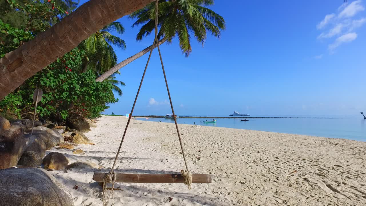 Travel concept with hanging seesaw on palm trunk bent over white sandy beach washed by blue sea in Thailand