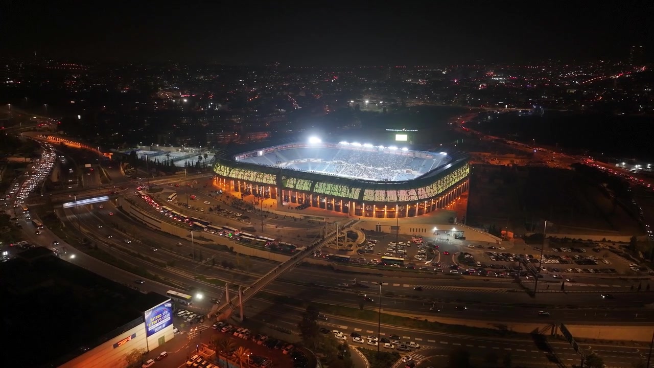 Prospettiva elevata dello Stadio Teddy illuminato di notte durante una partita con traffico autostradale circostante a Gerusalemme