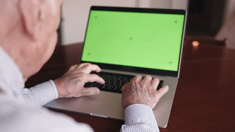 sobre el hombro del profesor sénior sentado en la mesa en la sala de estar y trabajando en una computadora portátil de moda con tecla de croma de pantalla verde. hombre anciano escribiendo con sus viejas manos en la computadora