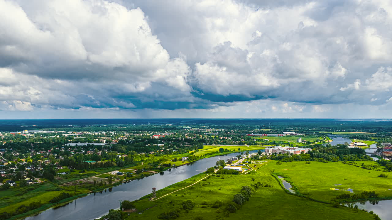 Aerial timelapse of a river winding through green fields and a small town under fast-moving dramatic clouds. Dynamic motion and changing light create an atmospheric landscape view