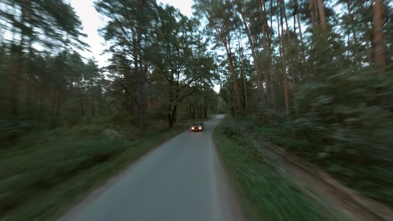 늦은 저녁에 옛 소나무 숲을 가로지르는 빈 은 시골 도로를 따라 속도를 내는 검은색 스포츠 자동차, fpv 레이싱 드론에서 촬영된 첫 번째 포인트 뷰