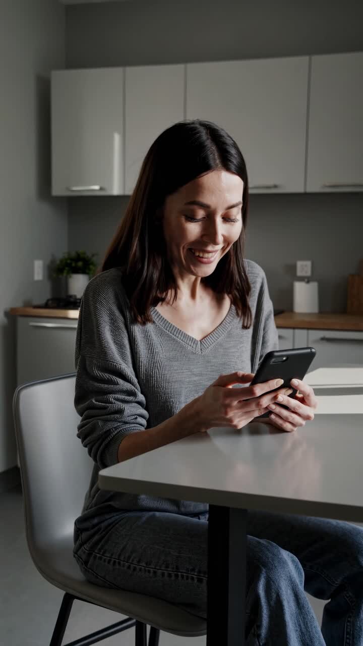 A woman in a gray sweater sits at a kitchen table, smiling at her phone