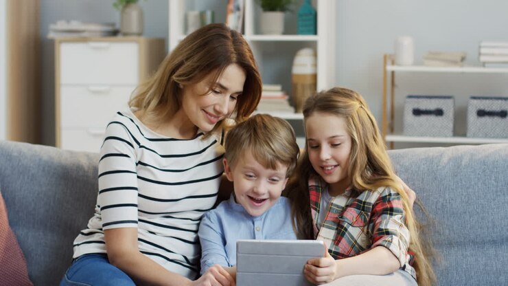 Portrait Of The Pretty Mother Sitting On The Gray Couch And Hugging Her Cute Small Son And Teen Daughter While They Watching Something On The Tablet Computer In The Living Room