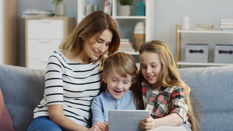Portrait-Of-The-Pretty-Mother-Sitting-On-The-Gray-Couch-And-Hugging-Her-Cute-Small-Son-And-Teen-Daughter-While-They-Watching-Something-On-The-Tablet-Computer-In-The-Living-Room