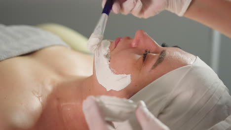 wellness technician applies white face scrub with silicone brush onto relaxed model wrapped in towel during spa treatment session amid soft studio lighting and professional salon environment