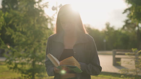 dama con ropa gris de pie al aire libre hojeando las páginas de un libro, mientras la luz del sol ilumina suavemente su figura y el fondo presenta árboles borrosos y un edificio